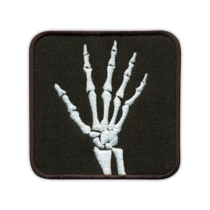 Op de afbeelding: Vierkante patch met een wit geborduurde skelet hand op een zwarte achtergrond. De patch heeft een bruine rand. De hand is in een gebaar van begroeting of afscheid, met de vingers licht gespreid.