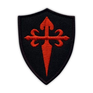May include: Black shield-shaped patch with a red cross of Santiago design. The cross features a sword-like vertical element and stylized arms. The patch has a black border and is likely made of fabric.