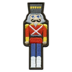 Könnte beinhalten: Gestickter Aufnäher eines Nussknacker-Soldaten. Der Aufnäher zeigt eine rote Jacke mit weißem Kreuz, blaue Hose und einen schwarzen Hut mit gelben Akzenten. Der Nussknacker hat einen weißen Schnurrbart und blaue Augen.
