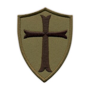 Pode incluir: Patch em forma de escudo verde-oliva com um desenho de cruz castanha. O patch tem uma borda costurada e uma aparência texturizada. A cruz está centrada no patch.