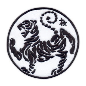 May include: Embroidered patch featuring a black tiger design within a white circle. The tiger is depicted in a dynamic pose, with intricate details. The patch has a black border and includes Japanese characters.