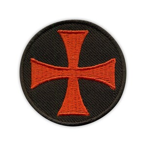 May include: Circular black patch featuring an embroidered orange cross. The cross has flared arms and a central vertical bar. The patch has a black border and is set against a white background.