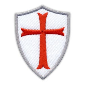 May include: White shield-shaped patch featuring a red cross design with flared ends. The patch has a grey border. It is likely an emblem or patch for clothing or accessories.