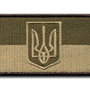 Könnte beinhalten: Gestickter Aufnäher mit dem ukrainischen Wappen, einem Dreizack, in Schildform. Der Aufnäher hat eine rechteckige Form mit einem dunkelbraunen Rand. Der Hintergrund ist in zwei horizontale Abschnitte aus Olivgrün und Beige unterteilt.