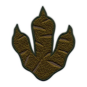 Op de afbeelding: Geborduurde patch in de vorm van een dinosaurusvoetafdruk. De patch heeft een gedetailleerd ontwerp met bruin- en groentinten en een donkergroene rand. De getextureerde uitstraling suggereert een hoogwaardig, duurzaam product, geschikt voor diverse toepassingen.