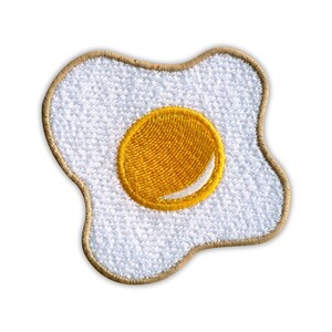 Op de afbeelding: Geborduurde patch met een gebakken ei ontwerp. Het eiwit is wit met een getextureerde uitstraling, en de dooier is heldergeel met een witte glans. De patch heeft een bruine rand en is geschikt voor kleding of accessoires.