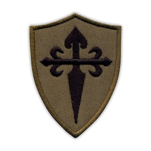 May include: A shield-shaped patch with a dark brown cross design on a tan background. The cross has a stylized arrow at the top and decorative flourishes on the horizontal arms. The patch is edged with a darker tan border.