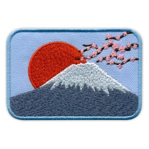 Peut inclure: Écusson brodé représentant le Mont Fuji avec un soleil rouge se levant derrière lui. La montagne est coiffée de blanc, avec une base bleu foncé. Des fleurs de cerisier roses sont dans le coin supérieur droit, sur fond bleu clair.