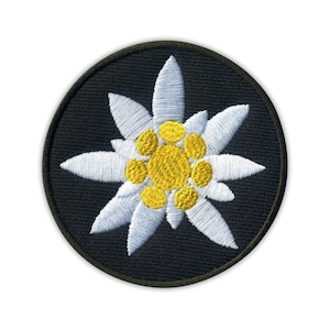 Puede incluir: Parche bordado con una flor de edelweiss blanca con centro amarillo sobre un fondo negro circular. El parche tiene un borde verde oscuro. Los pétalos de la flor están detallados con hilo blanco y el centro está relleno de amarillo.