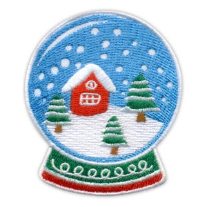 Könnte beinhalten: Ein runder Stickaufnäher mit einem Schneekugel-Motiv. Im Inneren befindet sich ein rotes Haus mit weißem Dach zwischen drei grünen Tannen auf weißem, verschneitem Boden, mit weißen Schneeflocken vor blauem Himmel. Der Sockel ist grün und rot.
