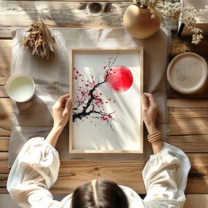 May include: A framed watercolor painting featuring a red sun and a black tree branch with red blossoms. The artwork is held by a person with a white blouse, set on a wooden table with other decorative items.