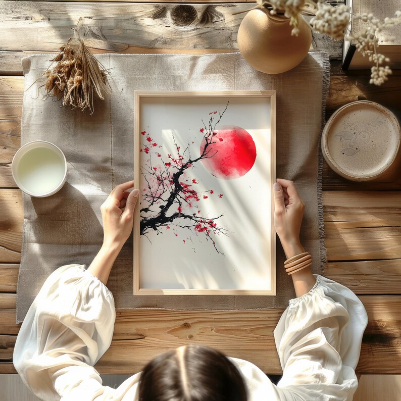 May include: A framed watercolor painting featuring a red sun and a black tree branch with red blossoms. The artwork is held by a person with a white blouse, set on a wooden table with other decorative items.