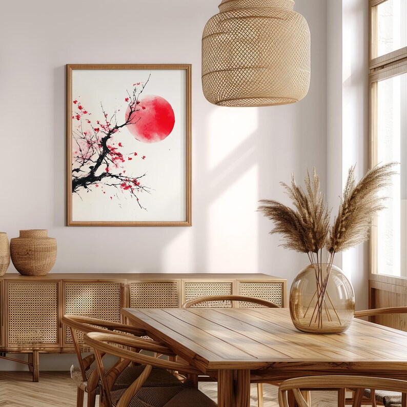 May include: A framed artwork featuring a red sun and a black tree branch with red blossoms. The artwork is in a wooden frame and hangs on a white wall. The room has a wooden table, chairs, and a woven light fixture.