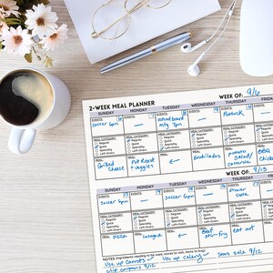 May include: A two-week meal planner with a gray and white design. The planner has a calendar layout with spaces for writing in meals for each day of the week. The planner includes a section for notes and a list of ingredients to use up. The planner is titled "2-Week Meal Planner" and includes the text "Week of: 9/16" and "Week of: 9/13".