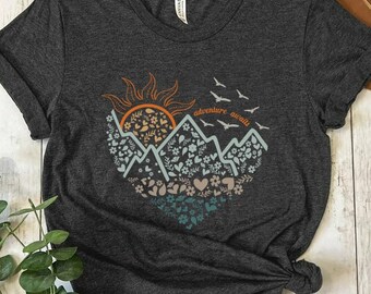 May include: Heather dark gray t-shirt with a graphic of a mountain range with a sun, birds, and flowers. The text "adventure awaits" is printed on the shirt.