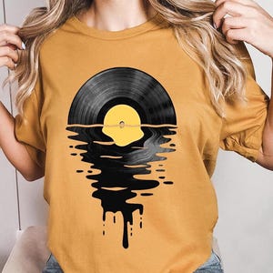 May include: A yellow t-shirt with a black vinyl record graphic that appears to be melting into the water. The record has a yellow center and is reflected in the water.