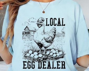 May include: Light blue t-shirt with a black and white graphic of a chicken, eggs, and a barn. The text on the shirt reads "LOCAL EGG DEALER". The shirt is made of a soft material and has a relaxed fit.
