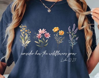 May include: Denim blue t-shirt featuring a floral design with the text "consider how the wildflowers grow" and the Bible verse Luke 12:27. The shirt has a relaxed fit and is made of a soft material.