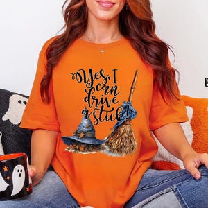 May include: A burnt orange short-sleeve t-shirt featuring a graphic of a blue witch hat and a broomstick with the black text Yes, I can drive a stick! The shirt is worn with blue jeans and is displayed alongside a black mug with white ghost designs.