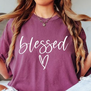 May include: A woman wearing a purple t-shirt with the word "Blessed" and a heart design in white. She is wearing blue jeans and has long blonde hair.