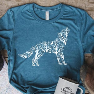May include: Teal t-shirt featuring a white wolf design made of tree branches and a crescent moon. A white mug with the text "And so the adventure begins" sits next to the shirt. Hiking boots and a backpack are also in the image.