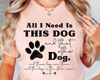 May include: Heather peach-colored t-shirt with the text "All I Need Is THIS DOG and that other Dog." A black paw print is centered below the text. The shirt is a casual style.