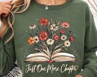 May include: A military green long-sleeve sweatshirt featuring a graphic of an open book with a bouquet of colorful wildflowers emerging from the pages. The text "Just One More Chapter" is printed in a white cursive font below the book illustration.