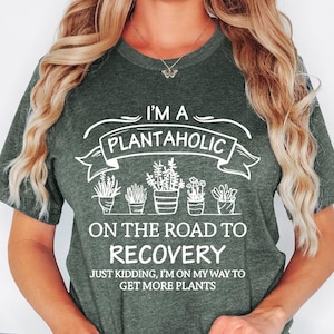 May include: A heather forest green short-sleeve t-shirt featuring white text that reads I'M A PLANTAHOLIC ON THE ROAD TO RECOVERY JUST KIDDING, I'M ON MY WAY TO GET MORE PLANTS, accompanied by a graphic illustration of five potted plants.