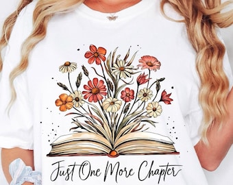 May include: A white t-shirt featuring a graphic illustration of an open book with a bouquet of colorful flowers emerging from the pages. Below the book, the text "Just One More Chapter" is printed in a black, cursive script font.