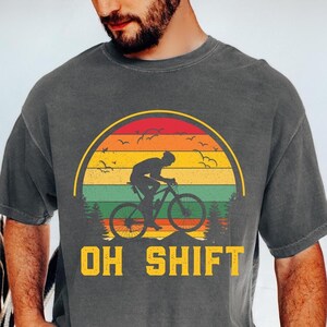 May include: A dark gray t-shirt with a vintage-style graphic of a person riding a bicycle in front of a sunset. The text "OH SHIFT" is printed below the graphic.