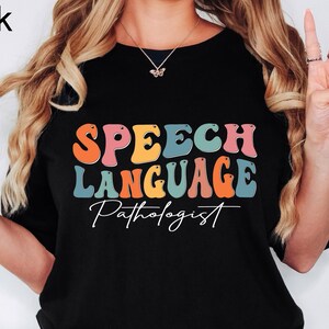 May include: Black t-shirt with the words "SPEECH LANGUAGE Pathologist" in colorful, retro-style lettering. The shirt is a crew neck and is made of a soft material.