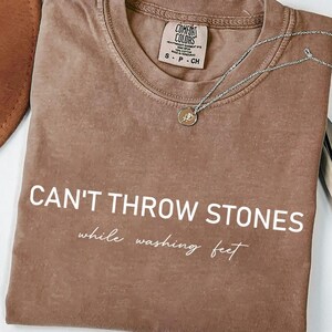 K&ouml;nnte beinhalten: Ein braunes T-Shirt mit dem Text "CAN'T THROW STONES while washing feet" in wei&szlig;en Buchstaben. Das Shirt ist gefaltet und liegt flach auf einer wei&szlig;en Oberfl&auml;che.
