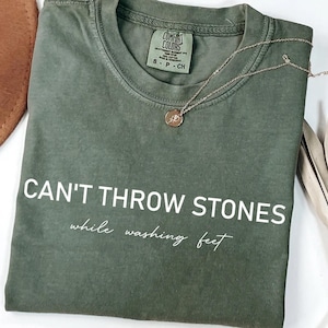 K&ouml;nnte beinhalten: Ein gr&uuml;nes Hanf-T-Shirt mit dem Aufdruck "CAN'T THROW STONES while washing feet" in wei&szlig; auf der Vorderseite.