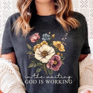 May include: A dark grey heather t-shirt with a floral design and the text "in the waiting GOD IS WORKING".