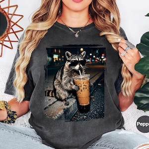 May include: A charcoal gray short-sleeve t-shirt featuring a graphic print of a raccoon wearing black sunglasses and holding a plastic cup of iced coffee. The raccoon is positioned on a curb at night with a blurred city street background.