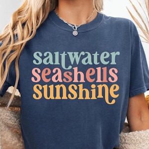 K&ouml;nnte beinhalten: Ein marineblaues T-Shirt mit den Worten "saltwater seashells sunshine" in Pastellfarben. Das Model tr&auml;gt Jeansshorts und eine cremefarbene Strickjacke. Ein dekorativer K&uuml;rbis und ein Weidenkorb befinden sich im Hintergrund.