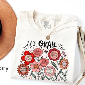 May include: An ivory short-sleeve t-shirt featuring a graphic of colorful flowers with text that reads: it's OKAY to make mistakes, start over, be different, have strong emotions, have bad days, ask for help, change your mind, and not know everything.