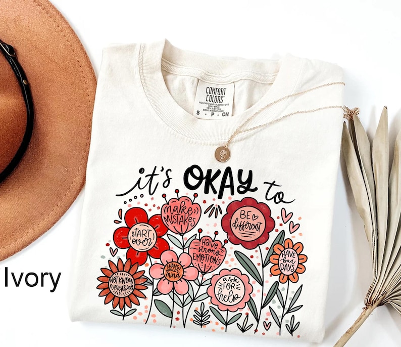 May include: An ivory short-sleeve t-shirt featuring a graphic of colorful flowers with text that reads: it's OKAY to make mistakes, start over, be different, have strong emotions, have bad days, ask for help, change your mind, and not know everything.