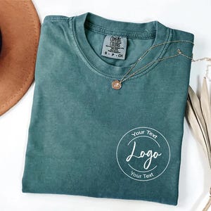 May include: A teal Comfort Colors t-shirt with a round neck and short sleeves. A gold necklace with a pendant rests on the shirt. A white circular logo with the words "Your Text" and "Logo" is printed on the front.