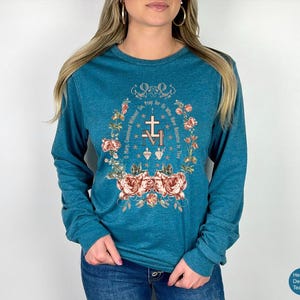 May include: Teal long-sleeve shirt with a floral and religious design. The design includes a cross, the letters "M" and "V", and the text "O Mary, Concerned Without Sin, Pray for Us Who Have Recourse to Thee."