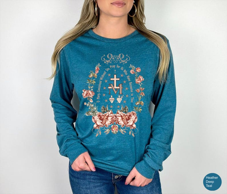 May include: Teal long-sleeve shirt with a floral and religious design. The design includes a cross, the letters "M" and "V", and the text "O Mary, Concerned Without Sin, Pray for Us Who Have Recourse to Thee."