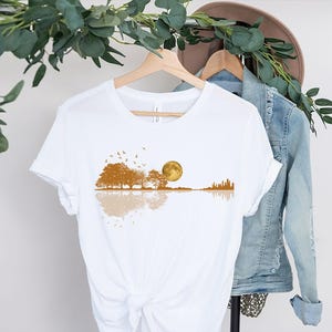 Pode incluir: Camiseta branca com um design em forma de guitarra que retrata uma cena de paisagem. O design inclui &aacute;rvores, uma lua cheia, p&aacute;ssaros e um horizonte da cidade em tons de marrom e dourado. O design &eacute; refletido abaixo da forma da guitarra.