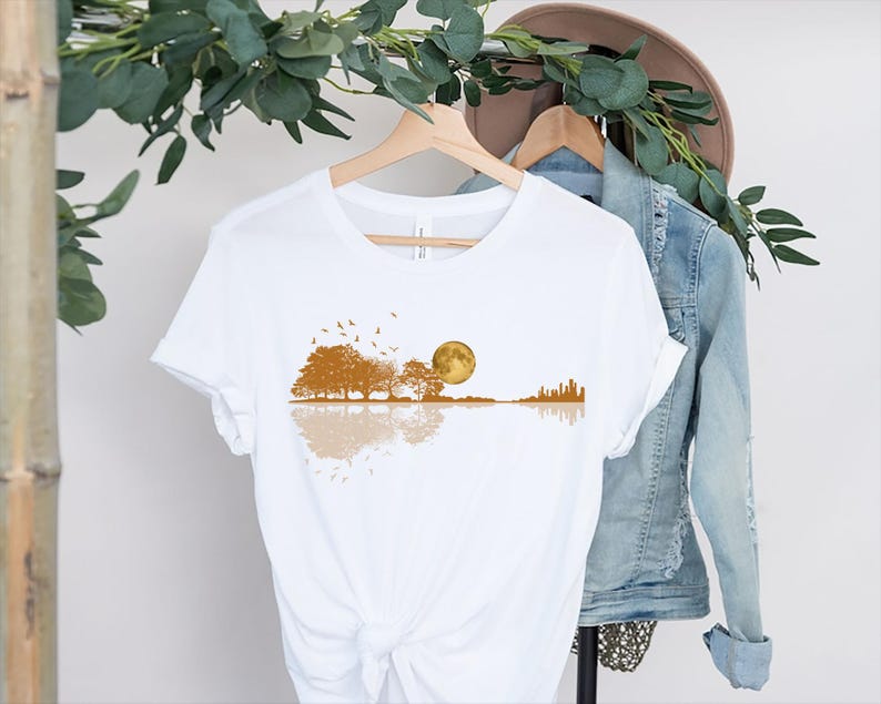 Pode incluir: Camiseta branca com um design em forma de guitarra que retrata uma cena de paisagem. O design inclui &aacute;rvores, uma lua cheia, p&aacute;ssaros e um horizonte da cidade em tons de marrom e dourado. O design &eacute; refletido abaixo da forma da guitarra.