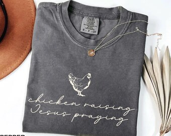 May include: A dark gray Comfort Colors t-shirt with a hen graphic and the words "chicken raising Jesus praying" in a cursive font. A silver necklace with a pendant is draped on the shirt. A brown hat is visible in the background.