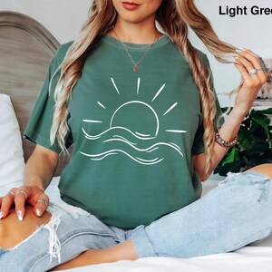 Pu&ograve; includere: Una t-shirt a maniche corte color verde chiaro con una grafica a linee bianche che raffigura un sole che sorge sopra le onde dell'oceano. Il testo "Light Green" &egrave; stampato nell'angolo in alto a destra. Ha un girocollo.