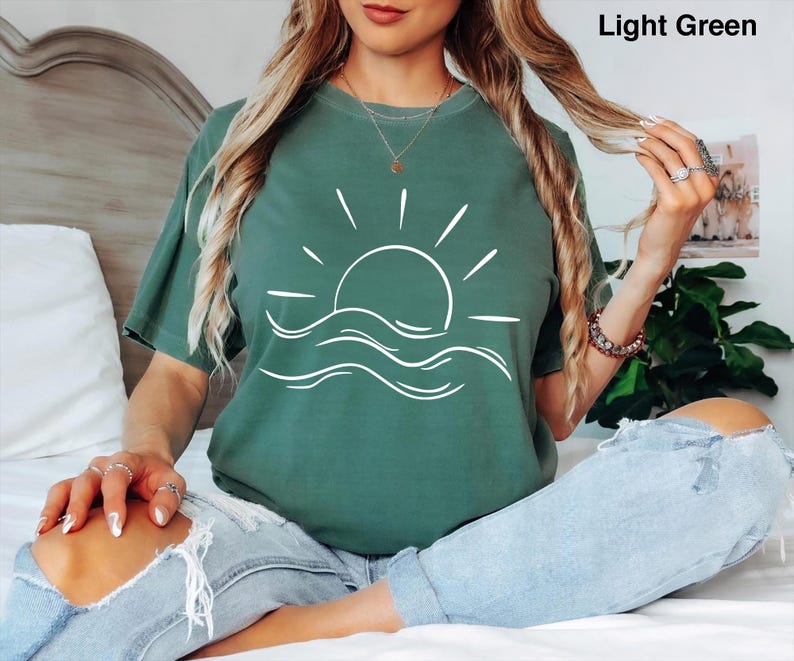 Pu&ograve; includere: Una t-shirt a maniche corte color verde chiaro con una grafica a linee bianche che raffigura un sole che sorge sopra le onde dell'oceano. Il testo "Light Green" &egrave; stampato nell'angolo in alto a destra. Ha un girocollo.