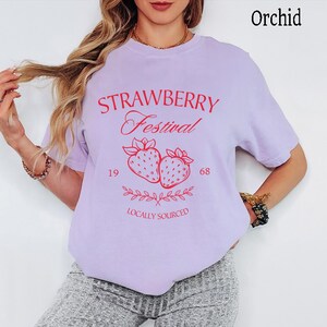 Op de afbeelding: Een orchideekleurig t-shirt met rode grafische tekst die STRAWBERRY Festival 19 68 LOCALLY SOURCED leest. Het ontwerp bevat een centrale illustratie van twee aardbeien met bebladerde steeltjes en een decoratief rankelement eronder.