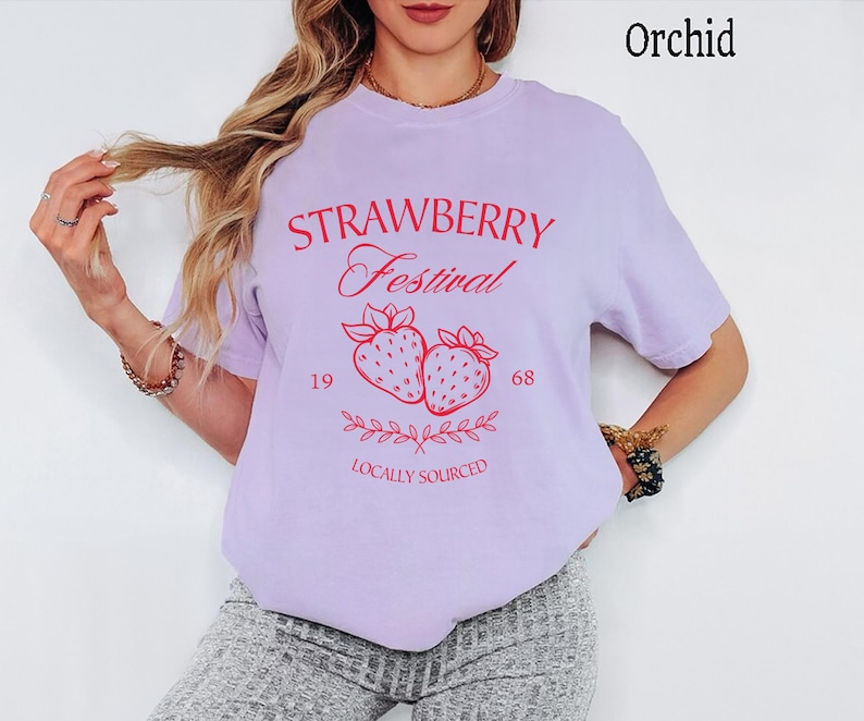 Op de afbeelding: Een orchideekleurig t-shirt met rode grafische tekst die STRAWBERRY Festival 19 68 LOCALLY SOURCED leest. Het ontwerp bevat een centrale illustratie van twee aardbeien met bebladerde steeltjes en een decoratief rankelement eronder.