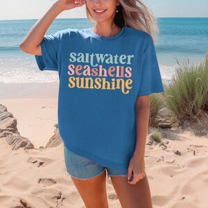 K&ouml;nnte beinhalten: Ein blaues T-Shirt mit dem Schriftzug "saltwater seashells sunshine" in einem farbenfrohen Retro-Schriftstil. Das Shirt wird am Strand getragen, mit Meer und Sand im Hintergrund. Es ist ein l&auml;ssiger, bequemer Stil.