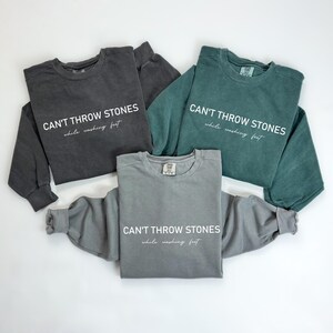 K&ouml;nnte beinhalten: Drei Sweatshirts in verschiedenen Farben, Grau, Schwarz und T&uuml;rkis, mit dem gleichen Text auf der Vorderseite: "CAN'T THROW STONES while washing feet".